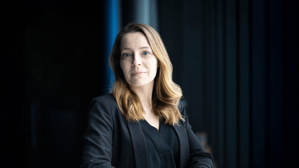 Ruut Kiiski seisoo tummasävyisessä tilassa pukeutuneena mustaan bleiseriin ja mustaan puseroon, valaistuna pehmeästi sivusta.