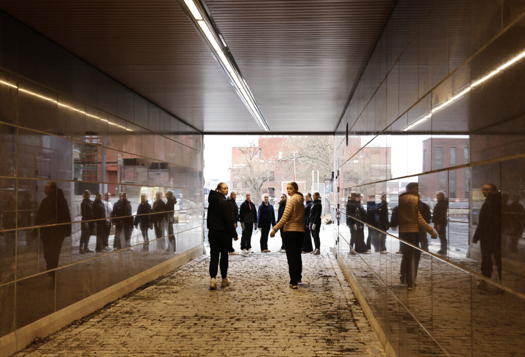 Alikulkutunneli, jossa kulkee ihmisiä ja heidän heijastuksensa näkyvät kiiltävillä seinillä.