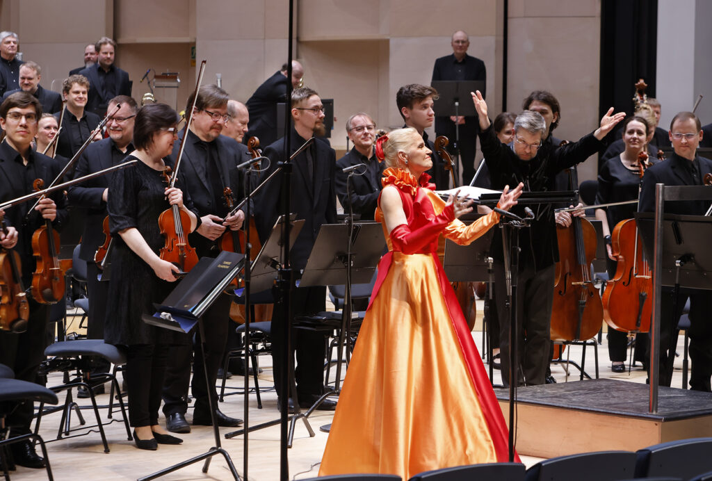 Sopraano Pia Komsi liekehtivässä oranssissa esiintymisasussa, taustalla Tampere Filharmonian orkesteri.