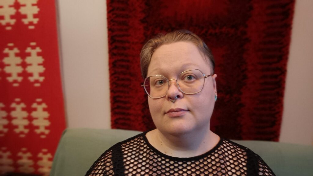 Alku Mattila mustassa reikäpaidassa, taustalla ryijy.