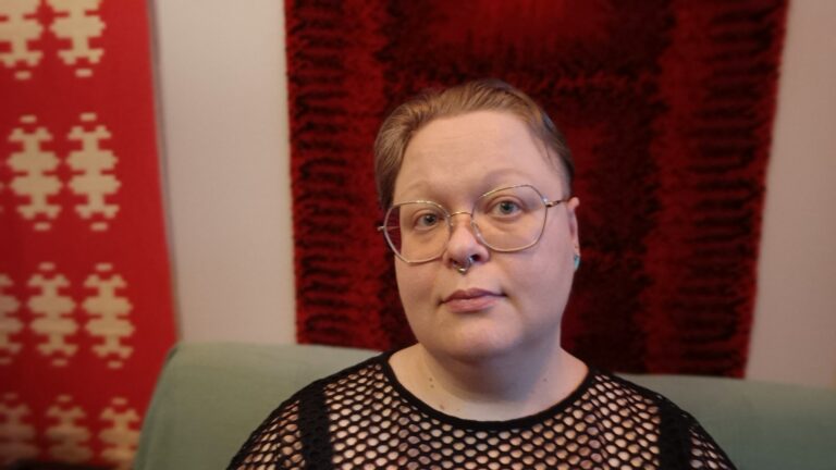 Alku Mattila mustassa reikäpaidassa, taustalla ryijy.