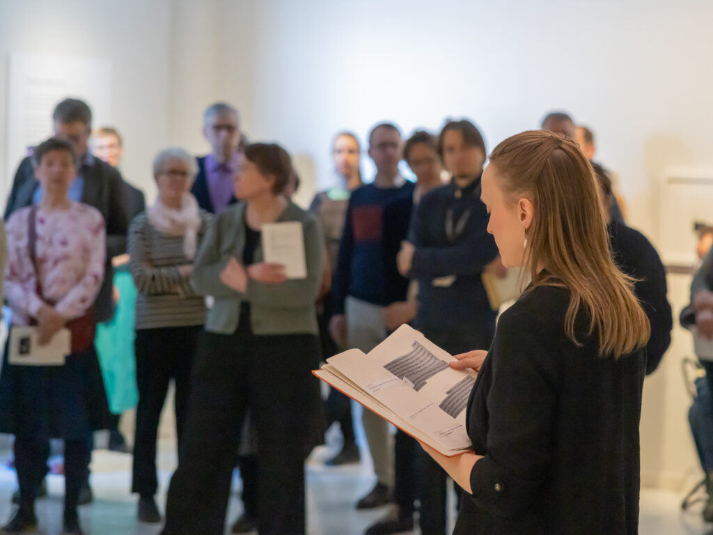 Ella Mettänen lukee tekstiä kirjasta, taustalla yleisöä.