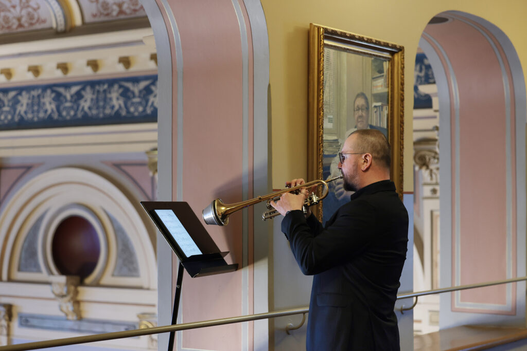 Trumpetisti soittaa parvella nuottitelineen edessä koristeellisen saliarkkitehtuurin keskellä.