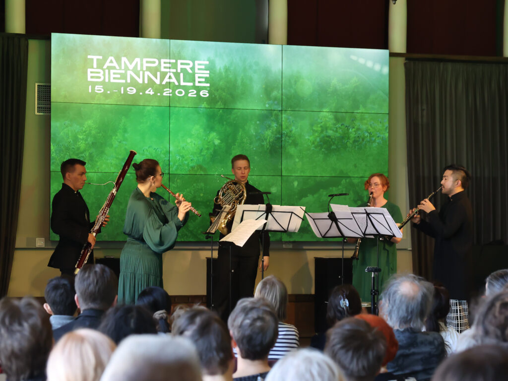 Laaja näkymä puhallinmusiikkiesityksestä lavalla ja yleisöstä etualalla.