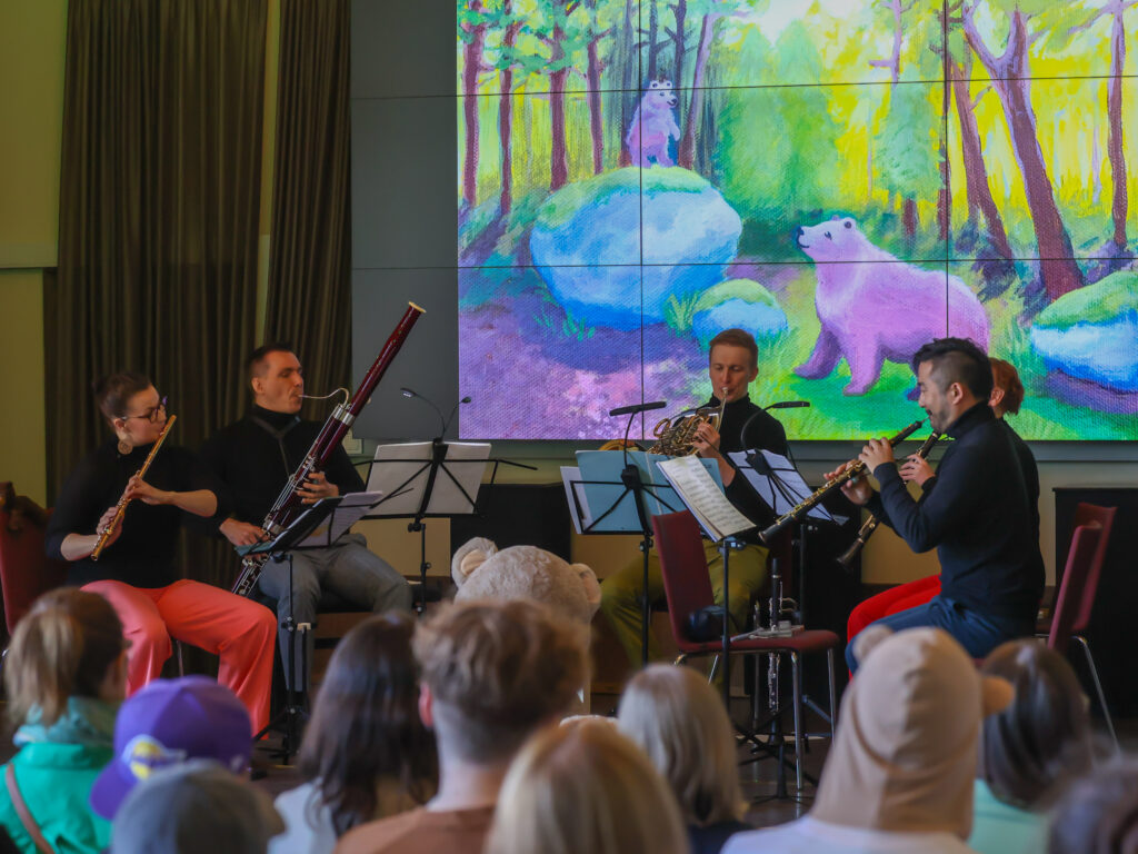 Puhallinmusiikki-esitys lapsiyleisölle, muusikot lavalla ja satumainen karhukuvitus taustanäytöllä.