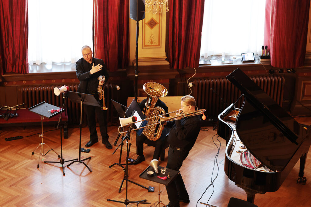 Vaskisoittajat esiintyvät flyygelin vieressä konserttisalissa, johtaja seisoo taustalla.