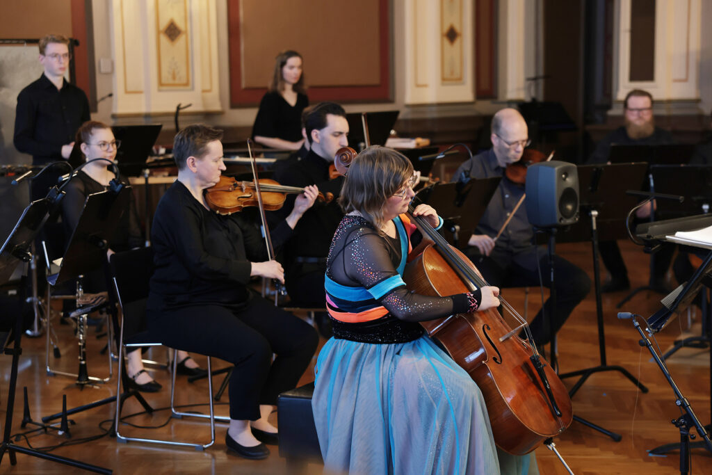 Sellisti soittaa orkesterin etualalla, taustalla jousisoittajia ja puhaltajia.